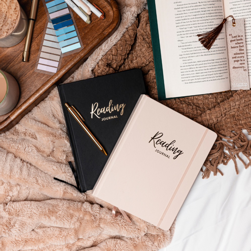 Two 'Reading Journal' notebooks on a textured surface with a book and coffee cups in the background.