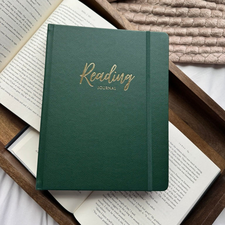 A dark green hardcover reading journal with gold foil lettering, styled on top of open books in a wooden tray with a chunky knit blanket in the background.